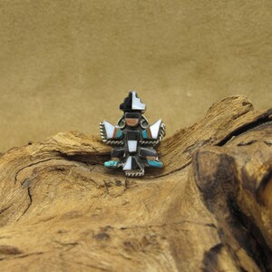 May include: A small, intricately crafted silver pendant featuring a geometric design inlaid with turquoise, black, white, and brown stones. The pendant is set against a rustic wooden background.