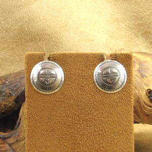 May include: A pair of round, silver-toned stud earrings. Each earring features a central design with a face-like motif, surrounded by a decorative border. The earrings are displayed on a brown, textured surface.