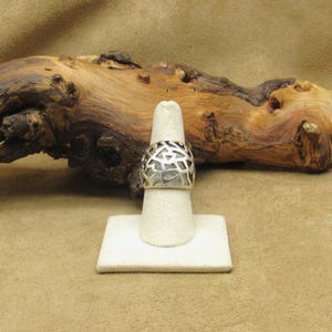 May include: A silver ring with an openwork design, displayed on a white ring holder. The ring's pattern features irregular shapes, and the background includes a piece of driftwood.