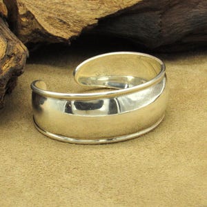 May include: A polished silver cuff bracelet with a curved design. The bracelet has a smooth, reflective surface and a rounded shape, with an opening to slip onto the wrist. The metal reflects the light, creating highlights and shadows.