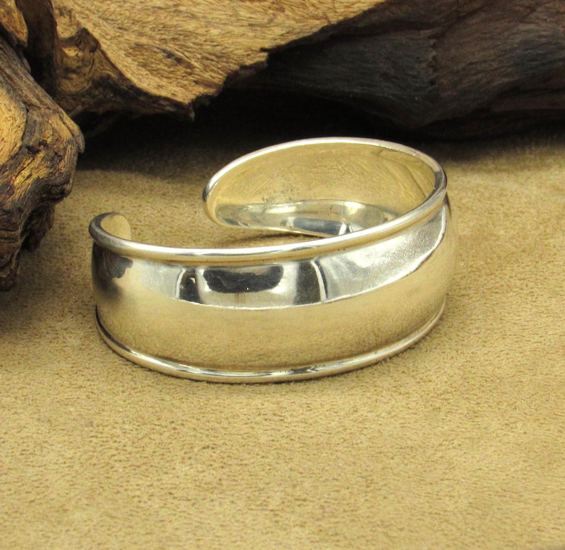 May include: A polished silver cuff bracelet with a curved design. The bracelet has a smooth, reflective surface and a rounded shape, with an opening to slip onto the wrist. The metal reflects the light, creating highlights and shadows.