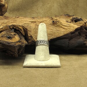 May include: A silver ring with an openwork design, displayed on a white ring holder. The ring features a repeating pattern of curved lines and small circles. The background includes a piece of driftwood and a neutral-toned surface.