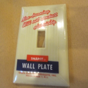 May include: Off-white Snapit wall plate with a rectangular opening for a light switch. The plate has vertical lines and text that reads "Non-burning, Will not conduct electricity." The bottom has a blue rectangle with "WALL PLATE" in white.