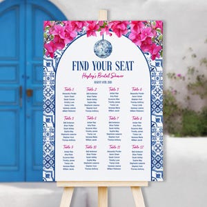 May include: A wedding seating chart on a wooden easel. The sign features a floral design with pink flowers and blue accents. The text "FIND YOUR SEAT" is displayed prominently, along with the bride's name and event date.