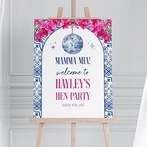 May include: A white welcome sign with a blue and white tile pattern border. The sign has pink flowers at the top and a disco ball hanging from an arch. The text reads "Mamma Mia! Welcome to Hayley's Hen Party August 17th, 2025".