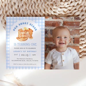 May include: A birthday invitation with a blue gingham border and the text "Our Short Stack Is Turning One." The invitation features a watercolor illustration of pancakes with butter and a photo of a smiling baby. The invitation includes the date, time, and location.