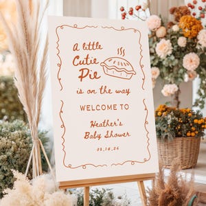 May include: A baby shower welcome sign on an easel. The sign has a cream background with brown text and a decorative border. The text reads "a little Cutie Pie is on the way WELCOME TO Heather's Baby Shower 08.14.26".