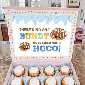May include: A pink box with a blue and white lid. The lid has a colorful graphic that says "There's no one Bundt you'd rather take to Hoco!" The box is filled with 12 small bundt cakes.