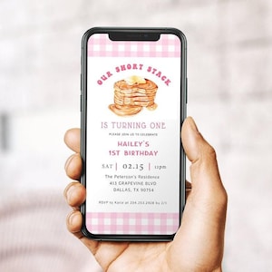 Puede incluir: Un teléfono inteligente muestra una invitación rosa y blanca a cuadros para un primer cumpleaños. La invitación presenta una pila de panqueques, el texto "Our Short Stack" y detalles de la fiesta.