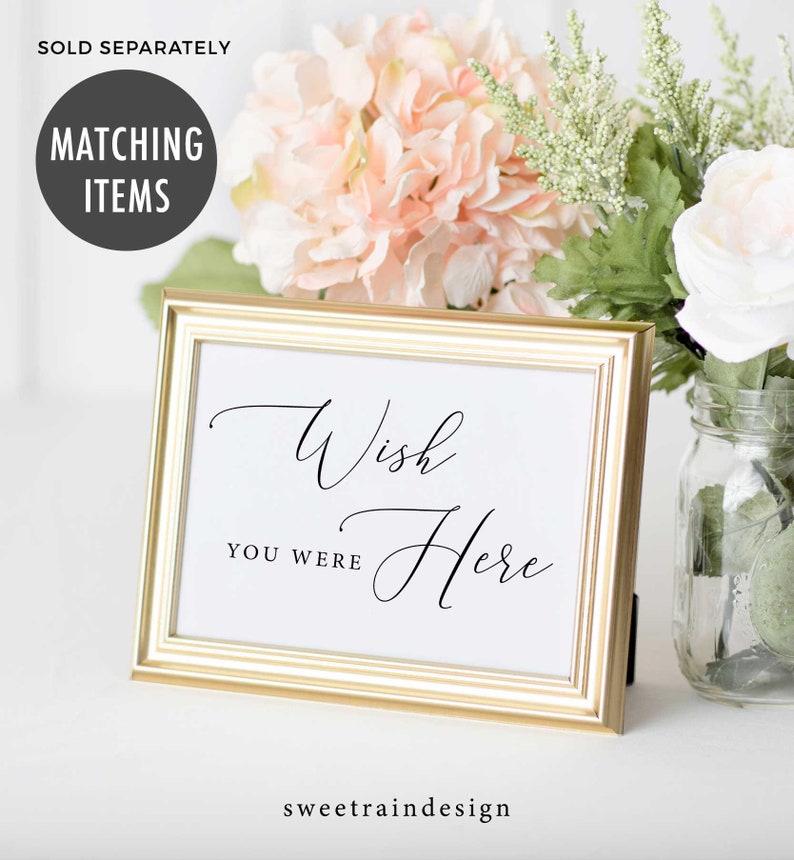 May include: A gold-framed sign with the text "Wish You Were Here" in a cursive font. The sign is sitting on a white surface with pink and white flowers in the background.