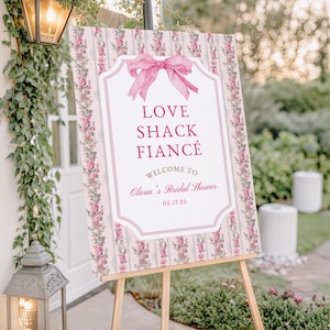 Puede incluir: Un letrero decorativo sobre un caballete de madera con el texto "LOVE SHACK FIANCÉ" y "Welcome to Olivia's Bridal Shower 05.17.25". El letrero tiene un lazo rosa y un patrón floral.
