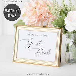 May include: A gold-framed sign with white paper inside. The sign says "Please sign our Guest Book" in black cursive font. The sign is sitting on a white surface with pink and white flowers in the background.