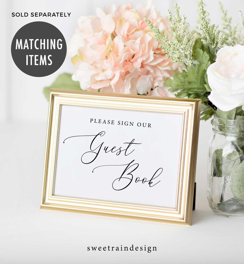 May include: A gold-framed sign with white paper inside. The sign says "Please sign our Guest Book" in black cursive font. The sign is sitting on a white surface with pink and white flowers in the background.