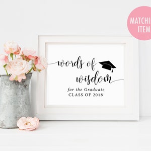 May include: White frame with a black and white sign that says "words of wisdom for the graduate class of 2018". A graduation cap is above the words. The sign is on a white surface with a bouquet of pink roses in a metal pitcher.