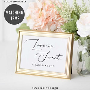 May include: Gold framed sign with the text "Love is Sweet" in black cursive font. Below the text is "Please Take One" in black font. The sign is sitting on a white surface with pink and white flowers in the background.