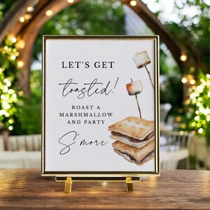 May include: A framed sign with the text "LET'S GET toasted! ROAST A MARSHMALLOW AND PARTY S'more." The sign features watercolor illustrations of toasted marshmallows on sticks and s'mores. The frame is gold and the sign is on a gold stand.