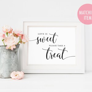 May include: White framed sign with black calligraphy that reads "Love is sweet, please take a treat". The sign is sitting on a white surface with a pitcher of pink flowers.