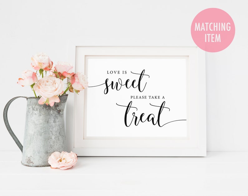 May include: White framed sign with black calligraphy that reads "Love is sweet, please take a treat". The sign is sitting on a white surface with a pitcher of pink flowers.