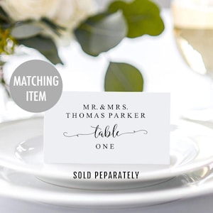 Op de afbeelding: Een wit naambordje met de tekst "MR. & MRS. THOMAS PARKER table ONE" in zwart schrift. De kaart ligt op een wit bord, met een vage achtergrond van groen en een wijnglas. Een grijze cirkel leest "MATCHING ITEM".