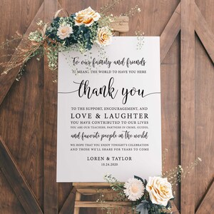Op de afbeelding: Een wit trouwbord met zwarte kalligrafietekst, waaronder "dank u" en een boodschap aan familie en vrienden. Het bord is versierd met bloemstukken en staat op een houten ezel tegen een houten deurachtergrond.
