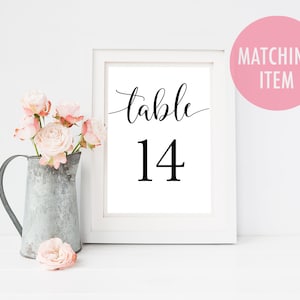 Op de afbeelding: Een ingelijste print met het nummer "tafel 14" in zwarte letters. De witte lijst staat op een wit oppervlak, met links een metalen kan gevuld met roze rozen. Een roze cirkel met de tekst "Matching Item" staat rechtsboven.