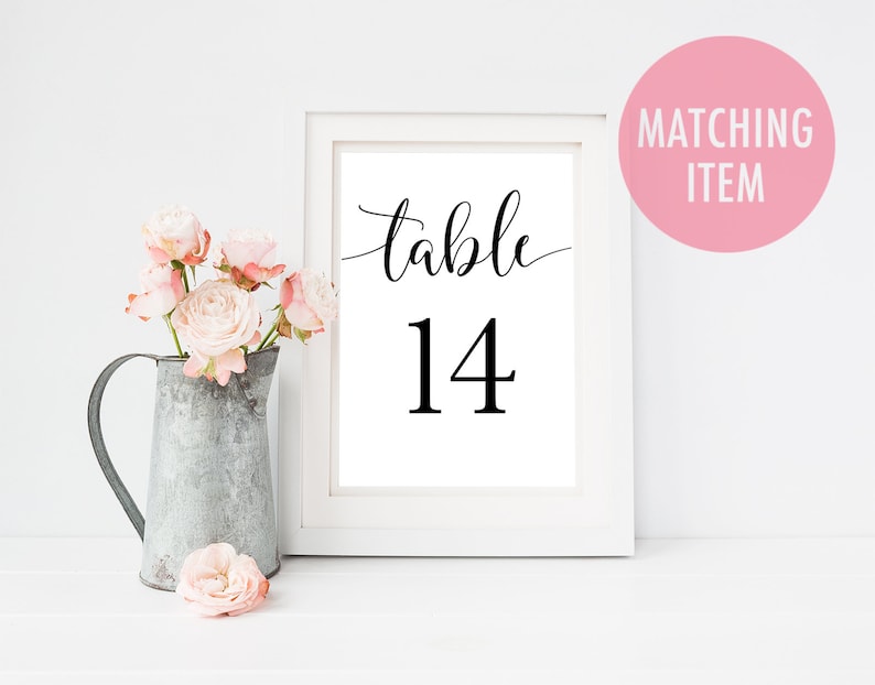 Op de afbeelding: Een ingelijste print met het nummer "tafel 14" in zwarte letters. De witte lijst staat op een wit oppervlak, met links een metalen kan gevuld met roze rozen. Een roze cirkel met de tekst "Matching Item" staat rechtsboven.