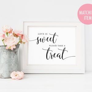May include: White framed sign with black calligraphy that reads "Love is sweet, please take a treat". The sign is sitting on a white surface with a pitcher of pink flowers.