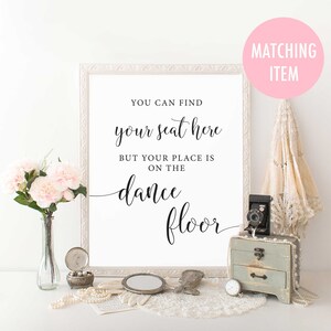 Puede incluir: Un cartel enmarcado en blanco con texto negro que dice "You can find your seat here but your place is on the dance floor". El cartel est&aacute; sobre una mesa con un mantel de encaje blanco, un jarr&oacute;n de flores rosas, una c&aacute;mara vintage y una peque&ntilde;a caja de madera.