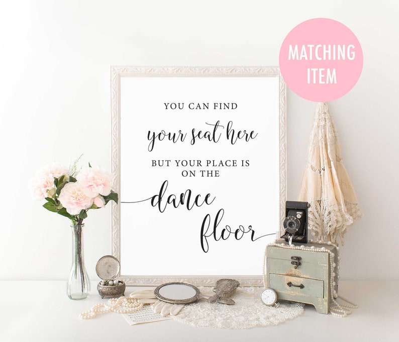 Puede incluir: Un cartel enmarcado en blanco con texto negro que dice "You can find your seat here but your place is on the dance floor". El cartel est&aacute; sobre una mesa con un mantel de encaje blanco, un jarr&oacute;n de flores rosas, una c&aacute;mara vintage y una peque&ntilde;a caja de madera.