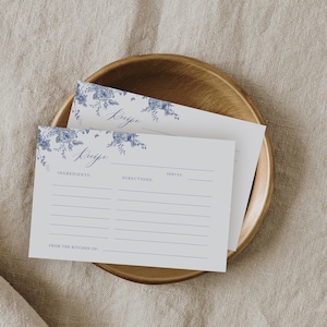 May include: Two white recipe cards with blue floral designs and the word "Recipe" written in cursive. The cards have lines for ingredients, directions, and servings. The cards are on a wooden plate.