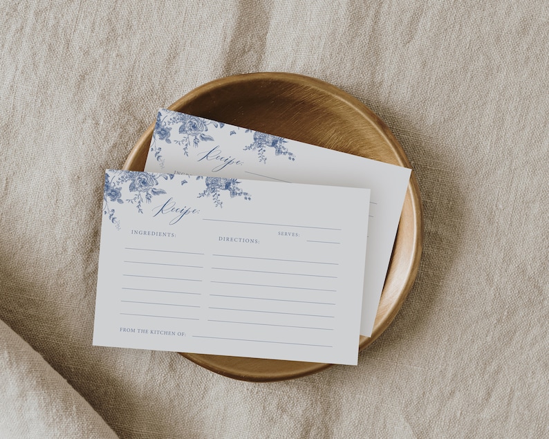 May include: Two white recipe cards with blue floral designs and the word "Recipe" written in cursive. The cards have lines for ingredients, directions, and servings. The cards are on a wooden plate.
