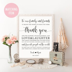 Op de afbeelding: Een ingelijst bord met elegante kalligrafie met de tekst "Aan onze familie en vrienden, bedankt". Het bord is omgeven door decoratieve items, waaronder bloemen, een vintage camera, een kleine kist en een parasol. De datum 06.14.2017 wordt ook weergegeven.