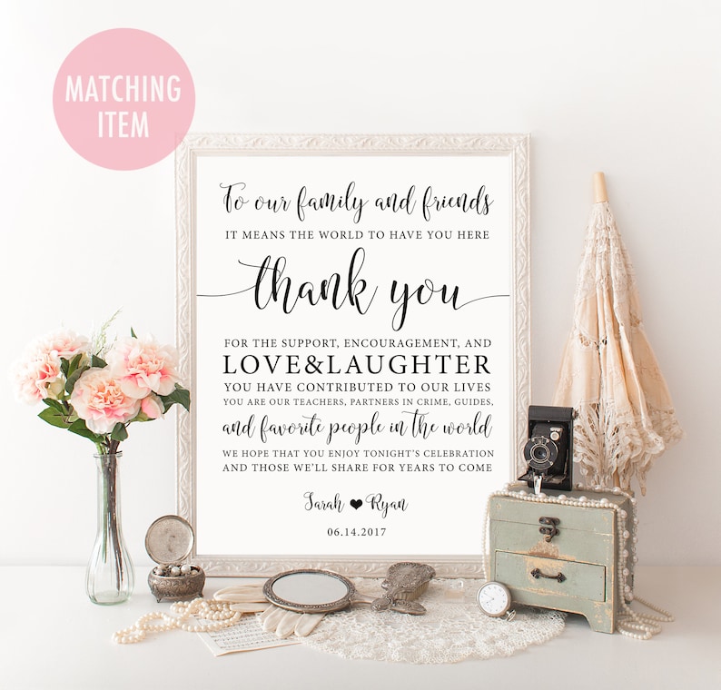 Op de afbeelding: Een ingelijst bord met elegante kalligrafie met de tekst "Aan onze familie en vrienden, bedankt". Het bord is omgeven door decoratieve items, waaronder bloemen, een vintage camera, een kleine kist en een parasol. De datum 06.14.2017 wordt ook weergegeven.