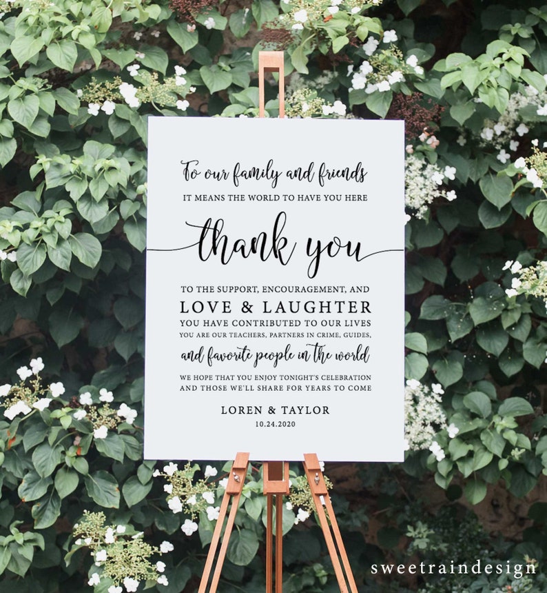 Op de afbeelding: Een wit bord op een houten ezel met de tekst "To our family and friends...thank you...Loren & Taylor 10.24.2020" in zwart schrift. Het bord is omgeven door groene bladeren en witte bloemen.
