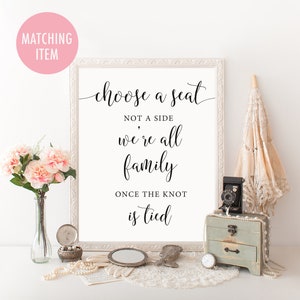Op de afbeelding: Een ingelijst bord met de tekst "choose a seat not a side we're all family once the knot is tied." Het bord is omgeven door decoratieve items, waaronder bloemen, een vintage camera en een kleine kist. Het bord staat in een witte lijst.