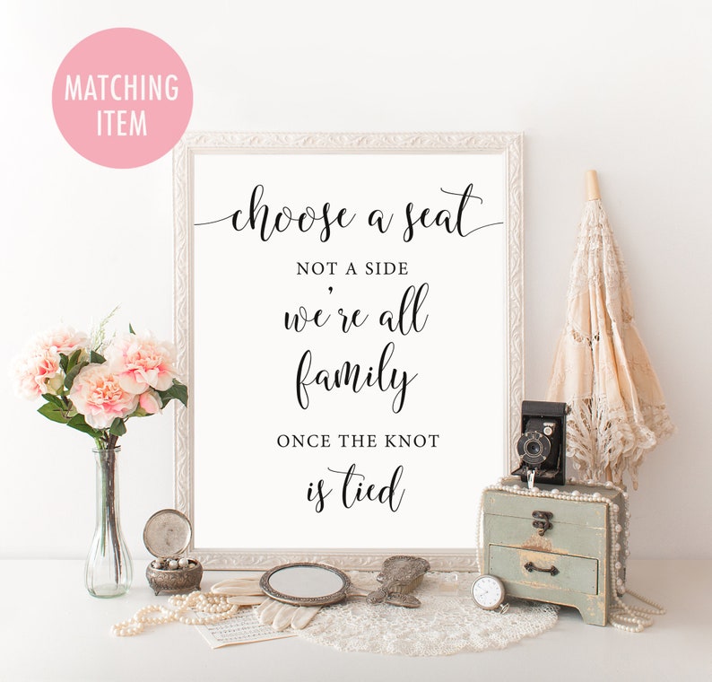 Op de afbeelding: Een ingelijst bord met de tekst "choose a seat not a side we're all family once the knot is tied." Het bord is omgeven door decoratieve items, waaronder bloemen, een vintage camera en een kleine kist. Het bord staat in een witte lijst.