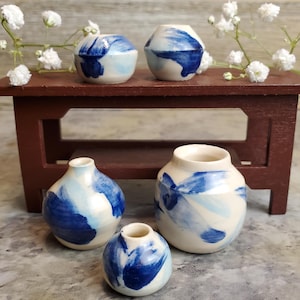 Puede incluir: Cuatro pequeños jarrones de cerámica con fondo blanco y pinceladas azules. Los jarrones están dispuestos sobre una mesa de madera.