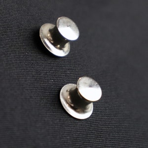 May include: Two silver metal hat-shaped push pins on a black fabric surface.
