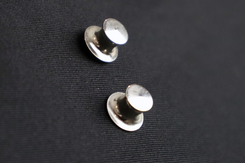 May include: Two silver metal hat-shaped push pins on a black fabric surface.