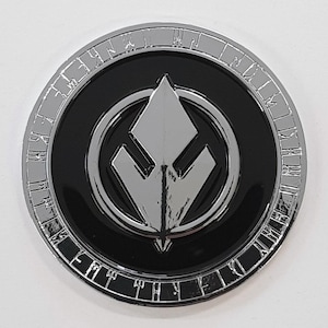 May include: A silver-coloured coin with a black centre featuring a stylised, silver-coloured symbol. The coin's edge is inscribed with a series of letters.