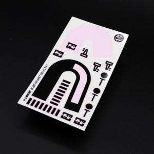 Peut inclure: Une feuille d'autocollants pour casque noirs et roses avec le texte "BOB-F HELMET DECALS V1" et un petit cercle avec un logo.