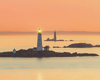 May include: Two lighthouses on rocky islands in the ocean at sunrise. The lighthouse on the left is lit and the light shines brightly. The sky is a soft orange color.
