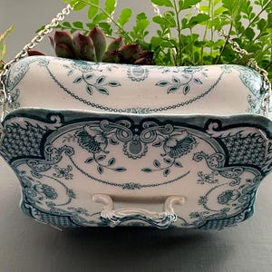 Op de afbeelding: Een hangende plantenbak van wit keramiek met een teal bloemenpatroon. De plantenbak wordt opgehangen door een zilveren ketting en is gevuld met groen en vetplanten. De plantenbak heeft een decoratieve, geschulpte rand.