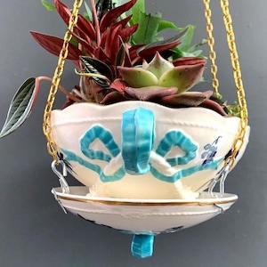 Op de afbeelding: Een hangende plantenbak met een witte keramische kom, versierd met blauwe lintontwerpen en een bijpassende handgreep. De plantenbak wordt opgehangen door een gouden ketting en bevat een verscheidenheid aan groene en rode planten. De schotel heeft een gouden rand.