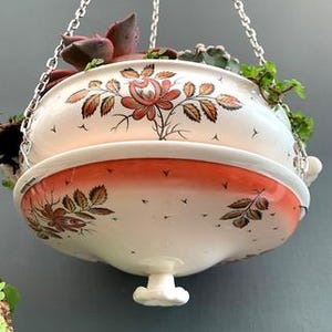 May include: A hanging ceramic planter with a floral design in shades of orange, brown, and white. The planter is suspended by a silver chain and filled with succulents and greenery. The planter has a decorative rim and a small, decorative foot.