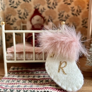 May include: A miniature white felt Christmas stocking featuring a fluffy pink faux fur cuff, a small gold star accent, and a glittery gold letter R embroidered on the front. The stocking is designed with visible stitching along the edges.