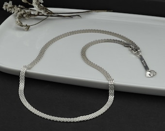 Könnte beinhalten: Eine silberne Kettenkette mit einem zarten, gewebten Design. Die Kette liegt auf einem weißen Teller mit einem kleinen Zweig weißer Blüten.