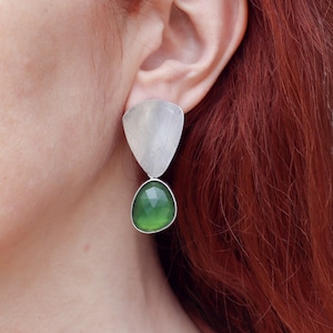 Könnte beinhalten: Silberne Ohrhänger mit einem grünen Edelstein. Die Ohrringe haben einen dreieckigen silbernen oberen Teil und einen tropfenförmigen grünen Edelstein.