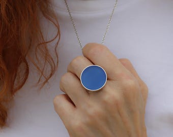 May include: A silver ring with a large, round, blue enamel center. The ring is worn on a finger, and a silver chain necklace is also visible. The background is a white shirt.