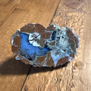 May include: A polished, oval-shaped stone with a striking pattern. The stone features a central area of light blue, surrounded by a white and brown outer layer. The surface has a cracked, textured appearance, showcasing natural geological formations.